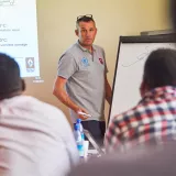 Man standing next to a white board talking to other people seated